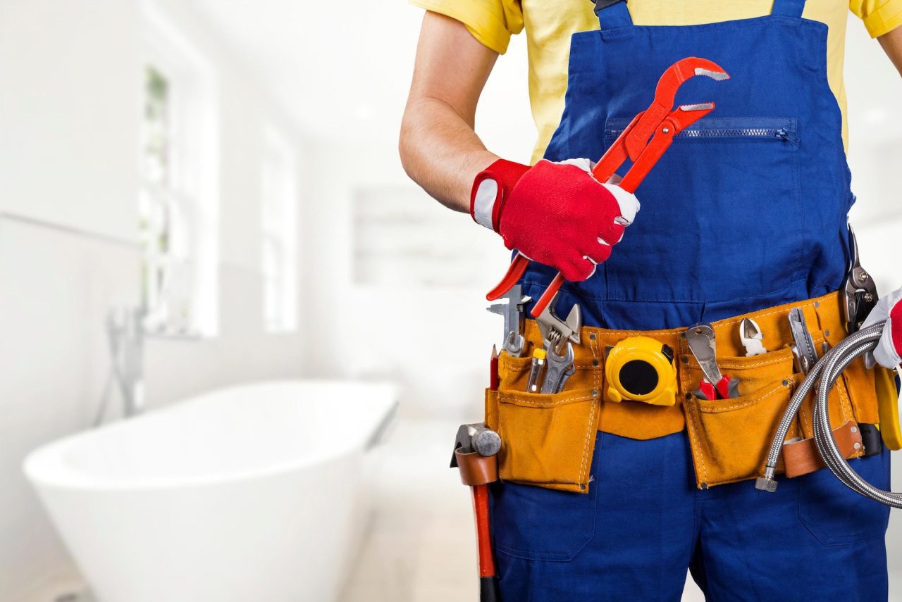 A plumber in a blue uniform holds a wrench, surrounded by tools, in a bright bathroom setting.